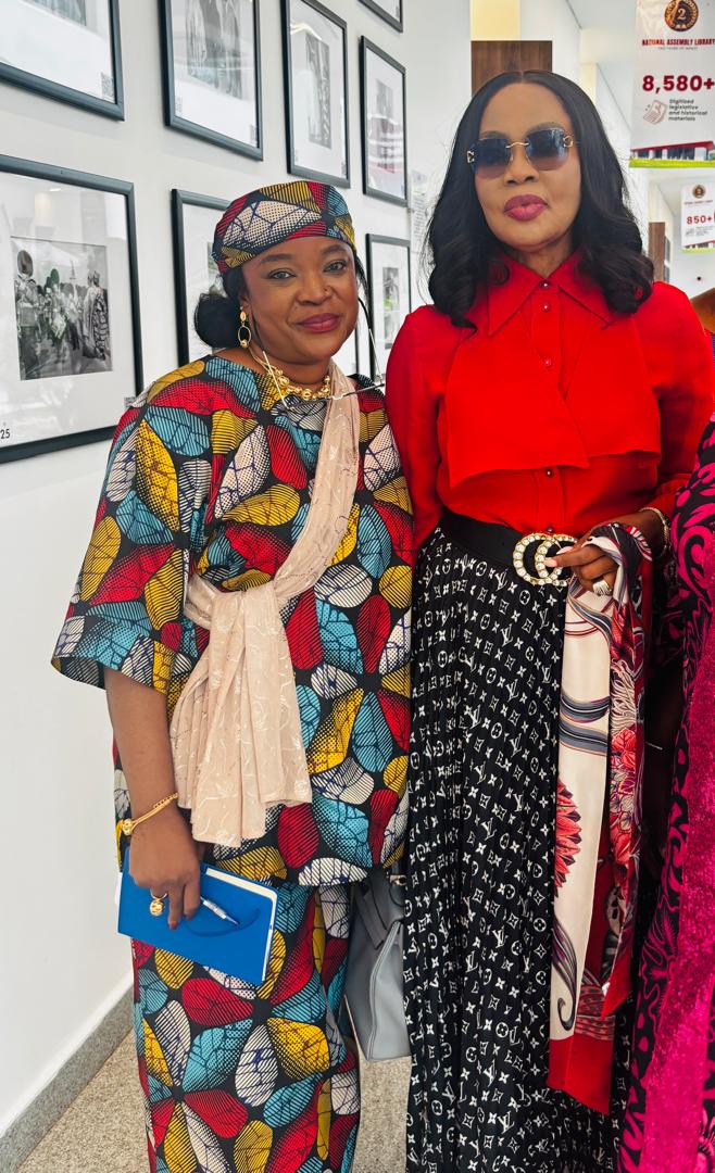 Anna Philemon Dara standing with Hon. Nkeiruka Onyejeocha beside a wall of framed photographs.
